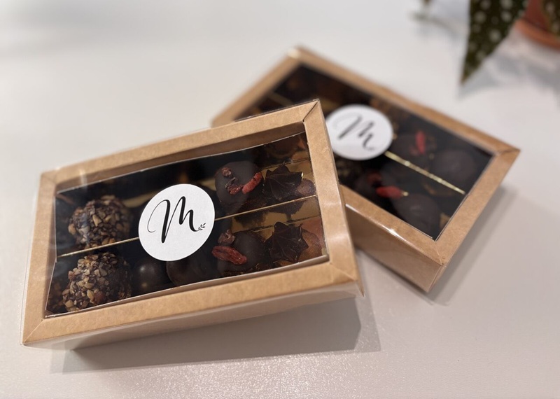 Due scatole in cartone kraft con finestra e bollino «M», piene di praline al cioccolato fondente con granella e bacche rosse
